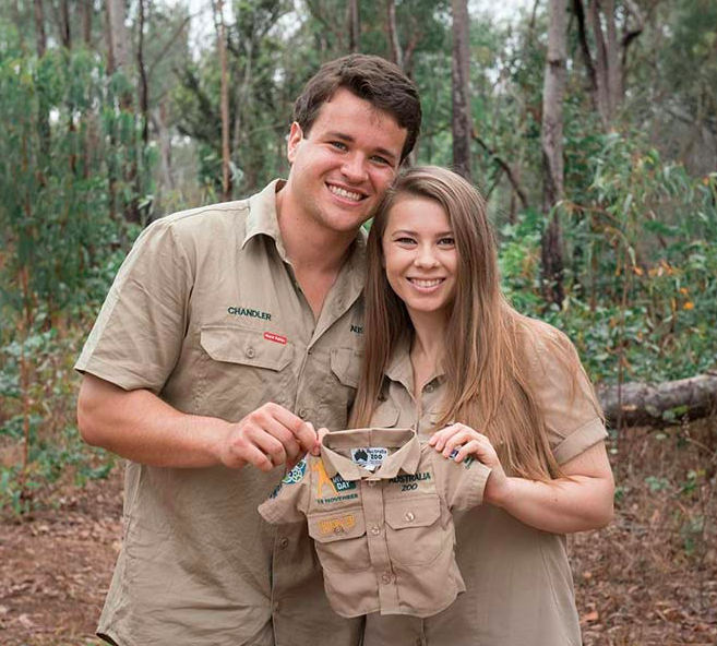 Couple Bindi Irwin and Chandler Powell expecting their first baby, holding up a little zoo keeper uniform.