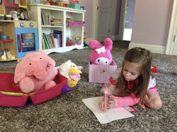 A young Hallie Zilberman writes on her bedroom floor in a pink cast, surrounded by pink stuffed animals. 